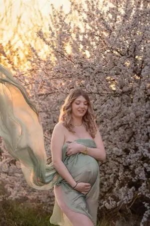 Femme enceinte nue avec un voile en extérieur devant un arbre fleuri.