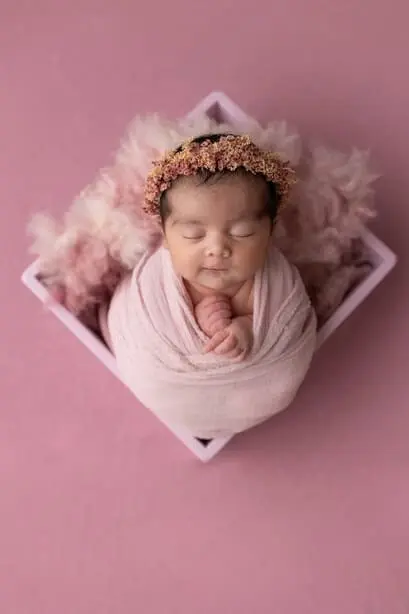 Bébé avec couronne de fleurs sur la tête et emmailloté sur fond rose.