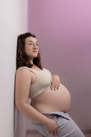 Femme enceinte de profil debout contre un mur, avec une main sur le ventre, dans les tons roses.