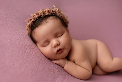 Bébé couché sur le ventre avec une couronne de fleurs sur la tête, tête posée sur les mains. Tons roses.